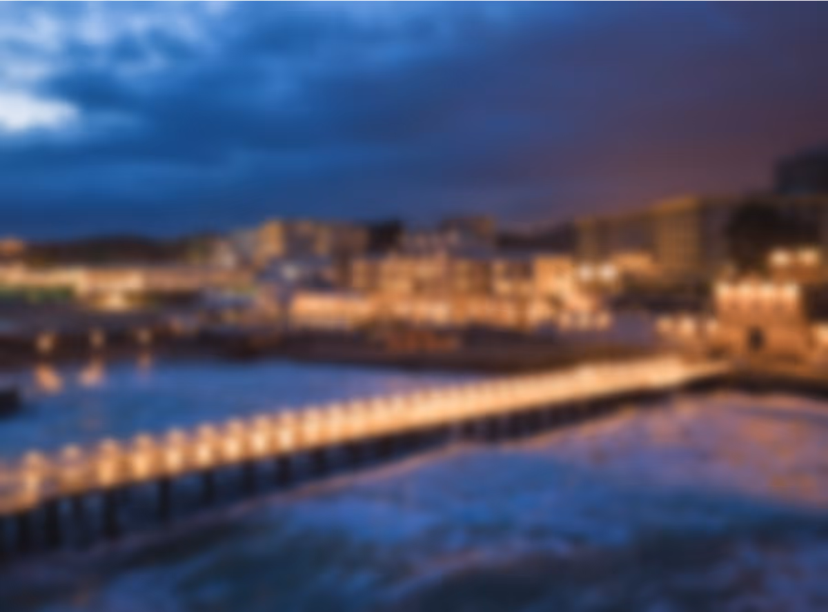 Blurry nighttime view of a lit pier extending over water with illuminated buildings in the background under a cloudy sky.
