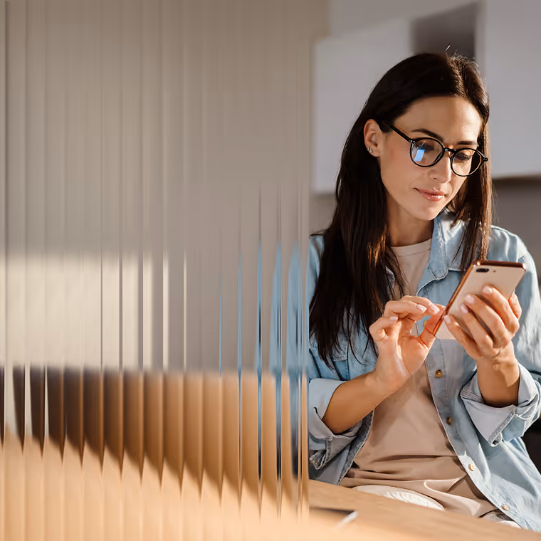 Woman with glasses using a smartphone while sitting at a table partially obscured by a vertical glass panel.