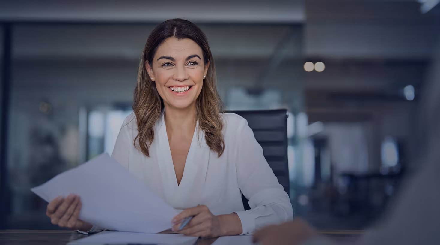Smiling woman in a white blouse holding documents in an office setting.