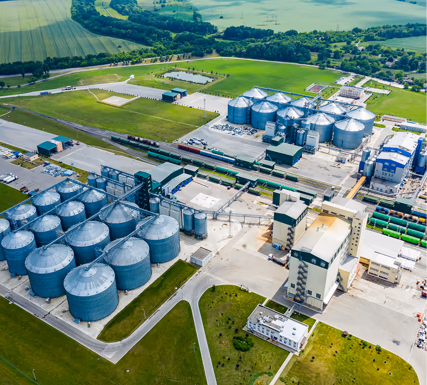 Aerial view of an industrial complex with multiple large silver silos, green fields, and train tracks with parked freight trains.