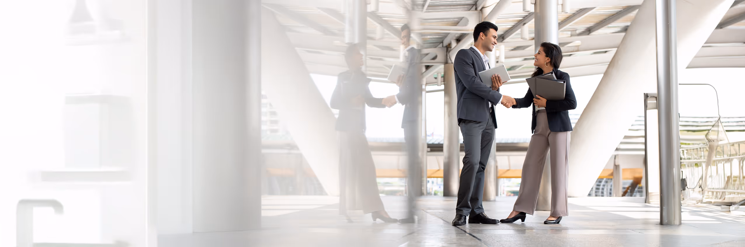 Two business professionals shaking hands in a modern office corridor, both holding folders and tablets.