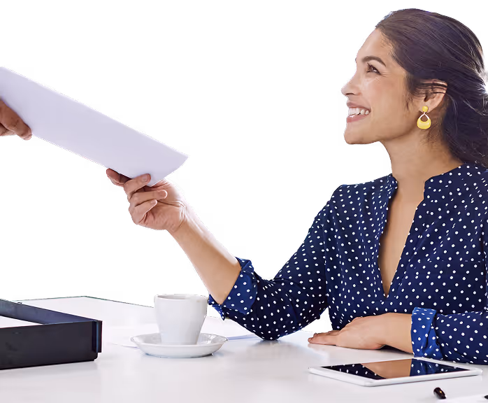Smiling woman in a blue polka dot blouse taking a document from another person at a desk with a coffee cup and tablet.