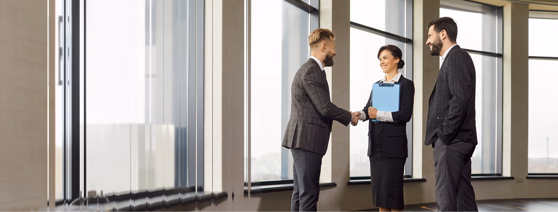 Two men and a woman in business attire shaking hands and smiling in a modern office with large windows.