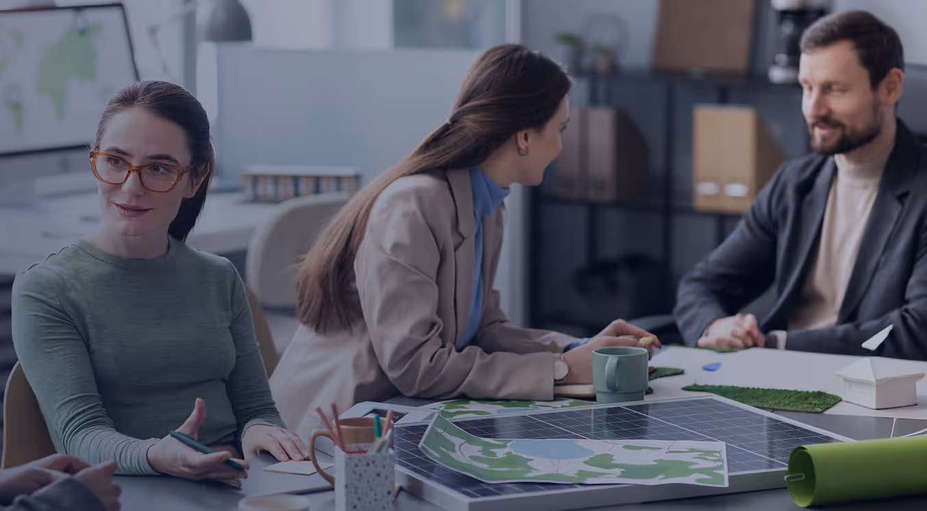 Three people sitting around a table discussing a solar panel model and maps in an office setting.