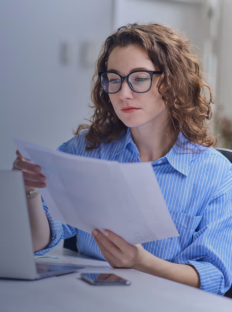 Woman wearing glasses and a blue striped shirt intently reading a document at a desk with a laptop and smartphone.