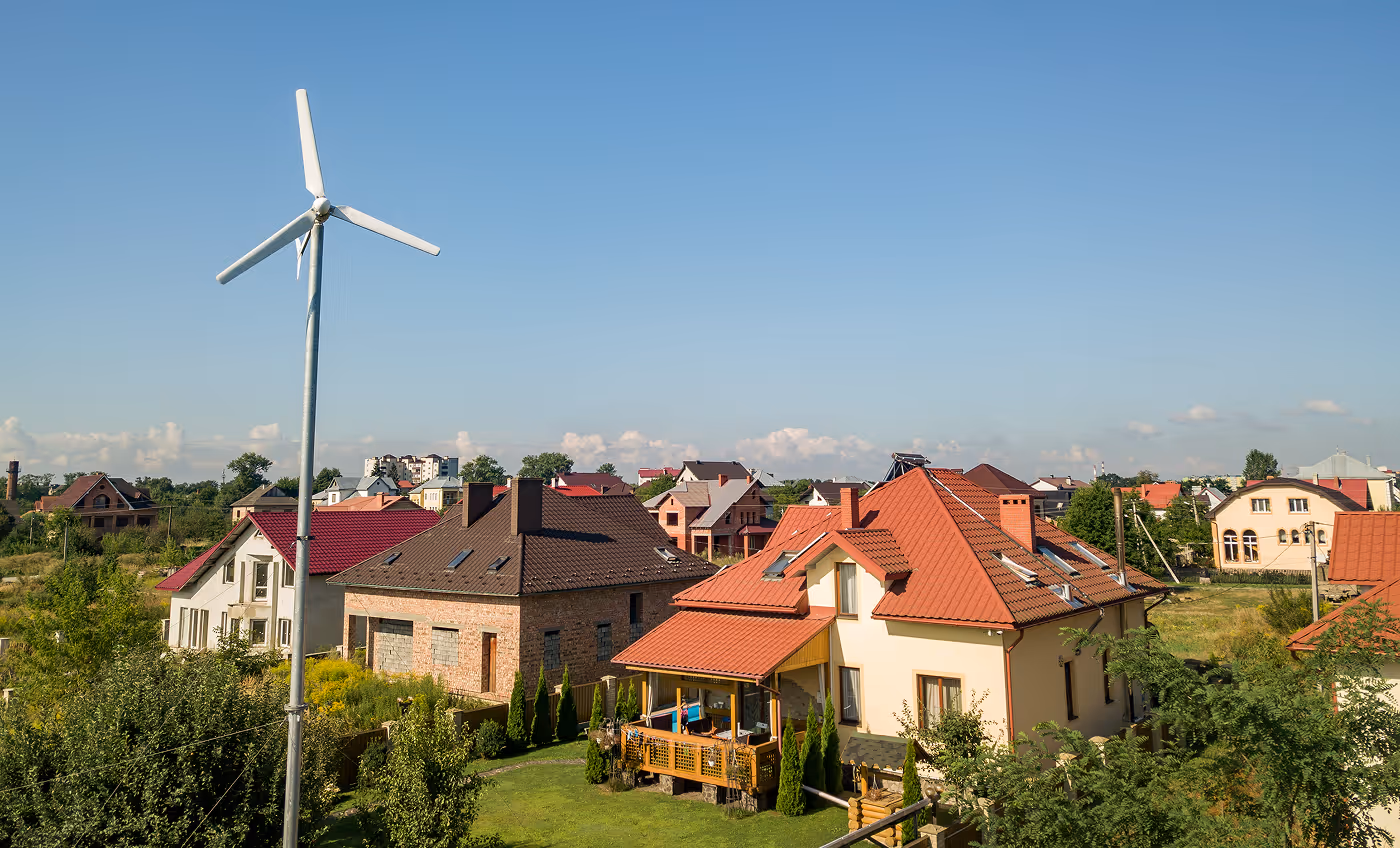 Residential neighborhood with a wind turbine and houses featuring red and dark brown roofs under a clear blue sky.