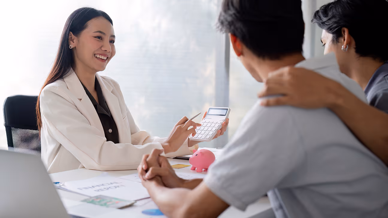 Smiling financial advisor using a calculator while talking to a couple seated at a desk with a pink piggy bank and documents.