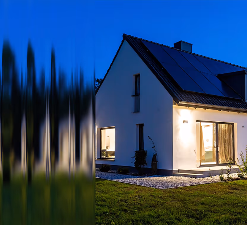 Modern house with solar panels on the roof during twilight, with warm indoor lights visible through windows.