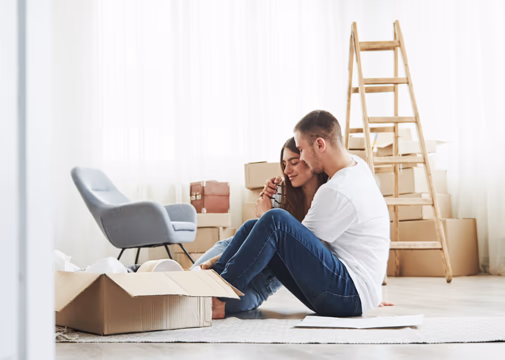 Couple sitting on the floor in a room surrounded by moving boxes and a wooden ladder, sharing a tender moment.