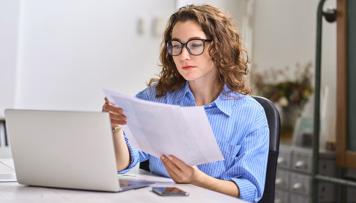 Woman with curly hair and glasses reviewing a document while sitting at a desk with a laptop and smartphone.