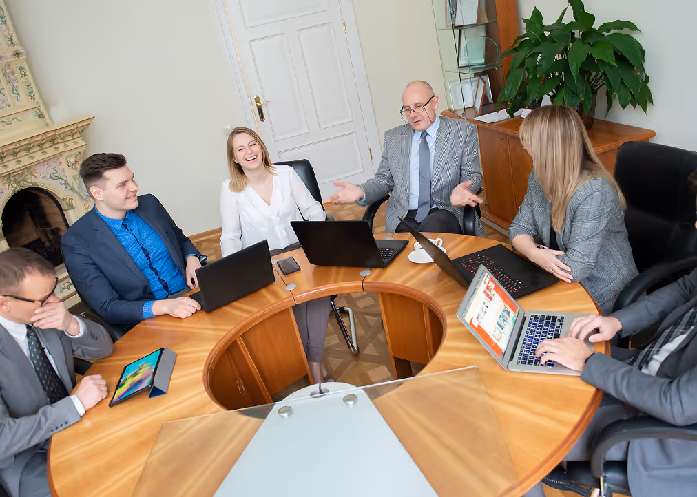 Five business people sitting around a round wooden table in an office, engaged in a discussion with laptops and a tablet.