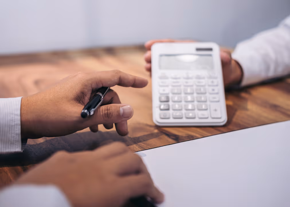 Two people at a wooden table, one holding a pen and the other holding a white calculator.