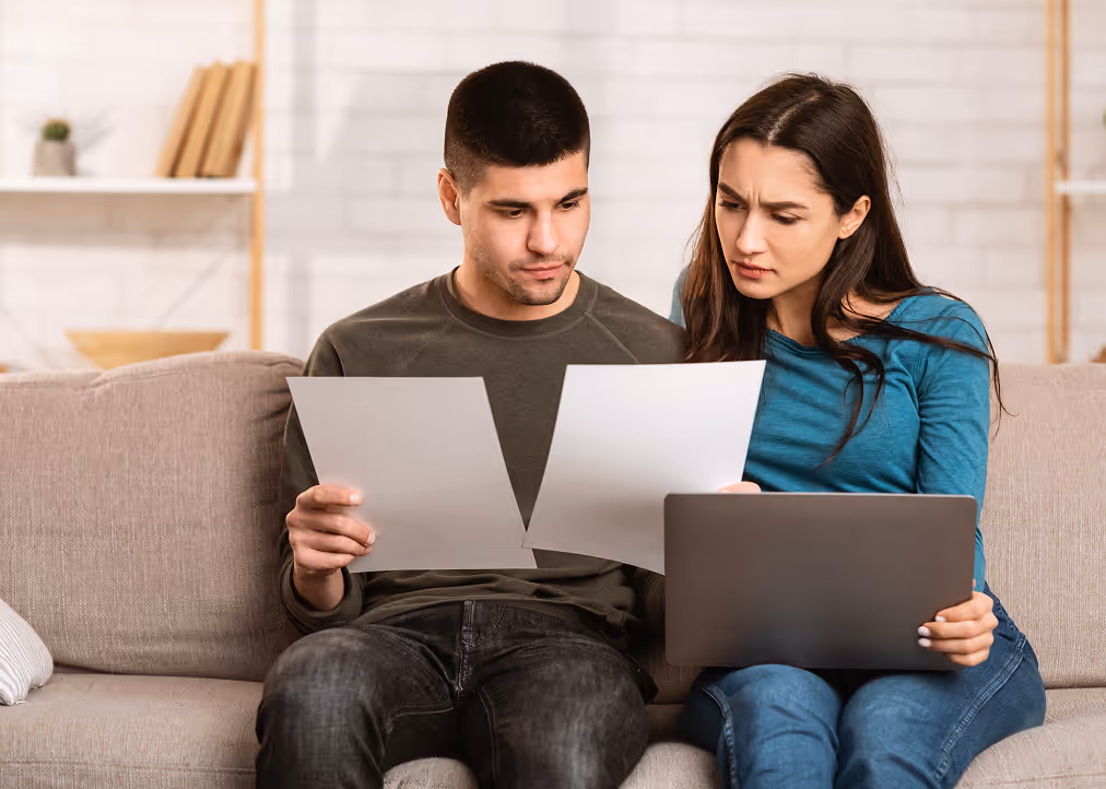 A man and woman sitting on a couch looking concerned while reviewing papers and a laptop.