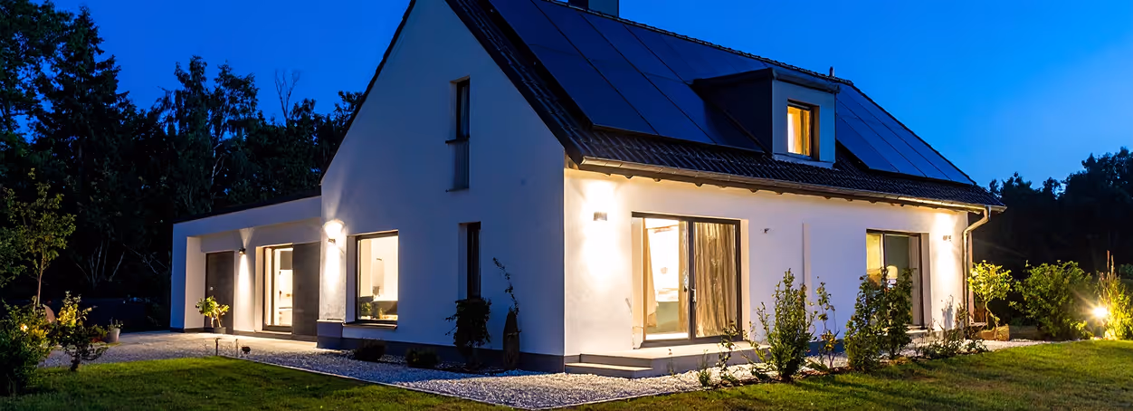 Modern white house with large windows and solar panels on the roof illuminated at twilight.