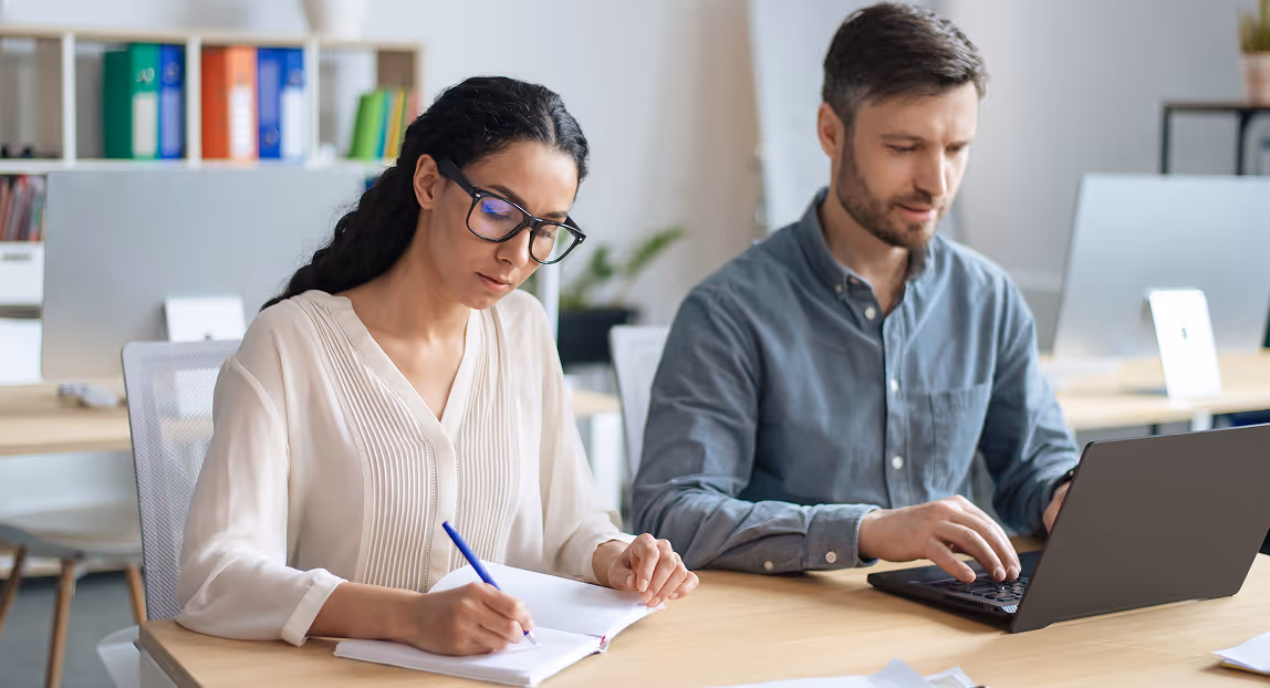 Two coworkers focused on their tasks, a woman writing in a notebook and a man typing on a laptop in a bright office.