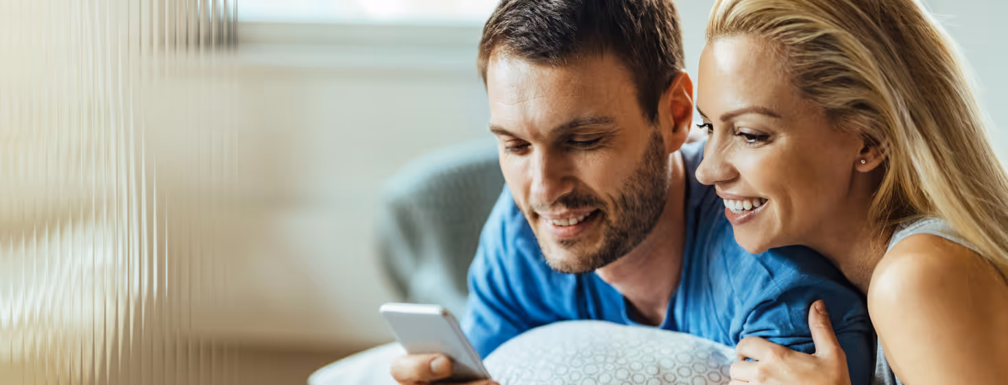 Smiling couple lying on a bed looking at a smartphone together.