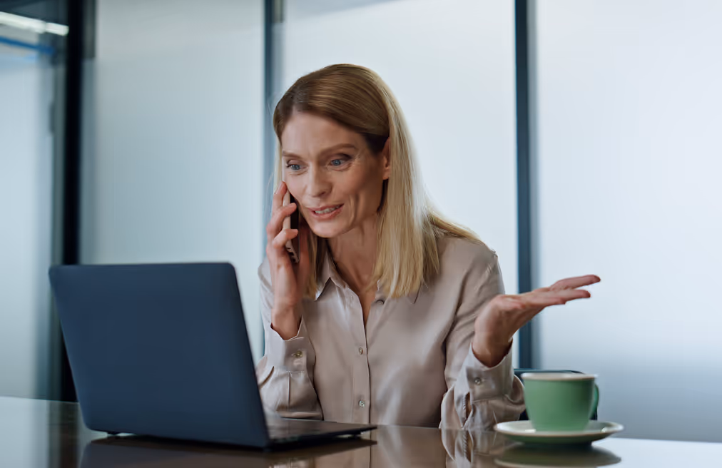 Woman talking on phone with a laptop open in front of her and a green coffee cup on the table.