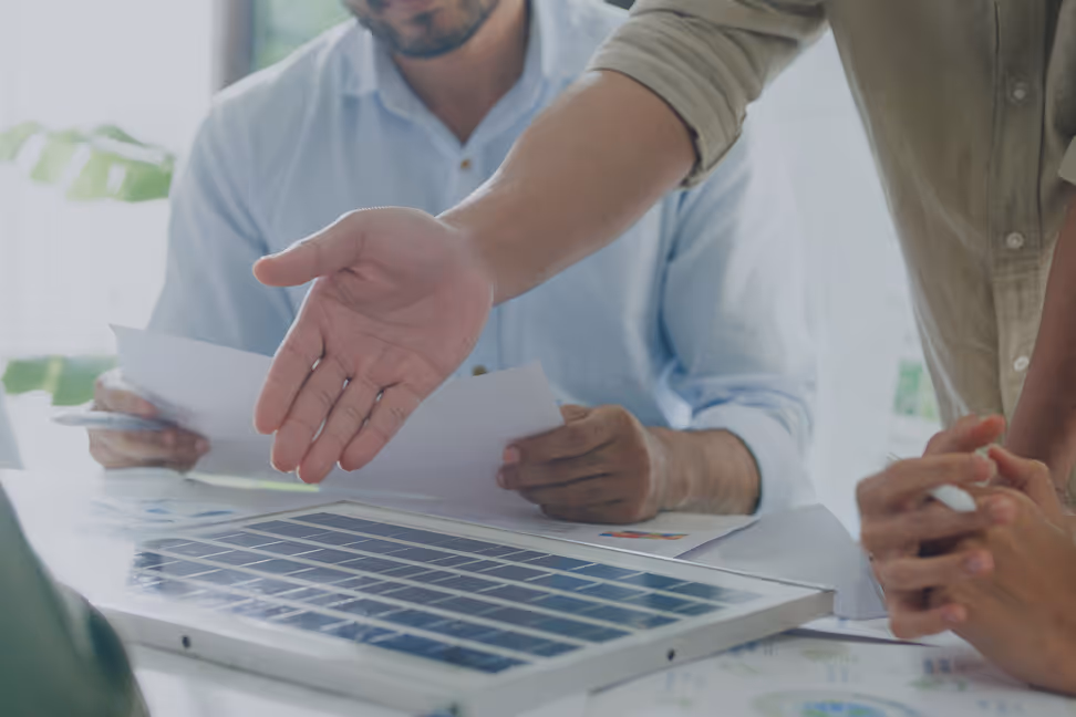 Person pointing at a solar panel on a table during a meeting with others reviewing documents.