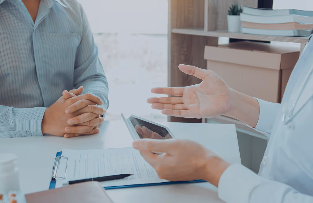 Doctor in white coat holding a tablet and explaining information to a patient sitting with folded hands at a desk.
