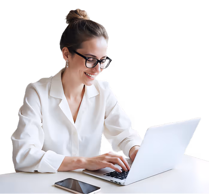 Smiling woman with glasses typing on a laptop at a white desk with a smartphone nearby.
