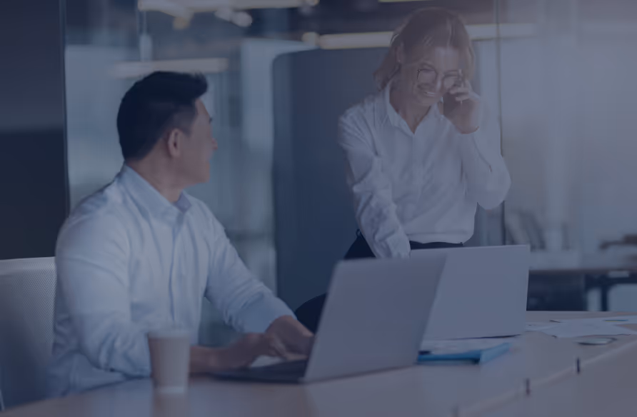 Two business professionals in a modern office, one sitting at a desk with a laptop and coffee cup, the other standing and talking on a phone.