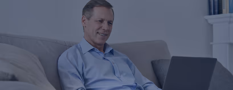Middle-aged man in a blue shirt sitting on a couch and looking at a laptop screen, smiling slightly.