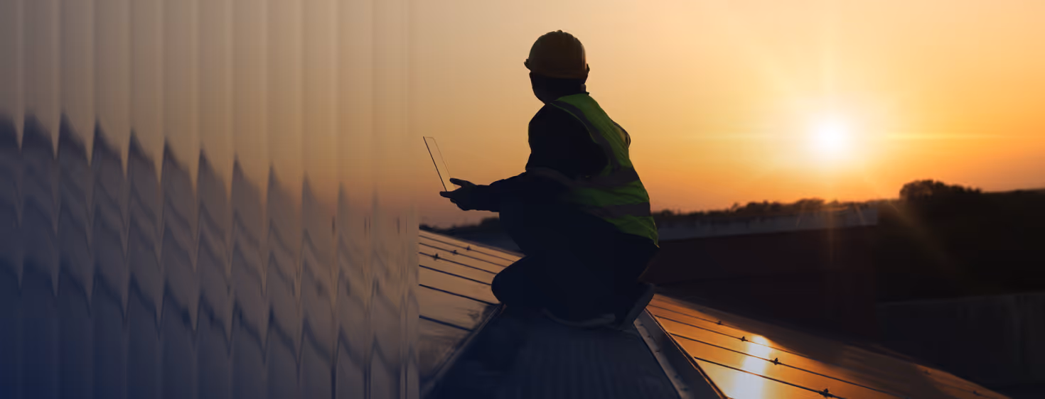 Silhouette of a worker in a safety vest and helmet crouching on a rooftop with a tablet at sunset.