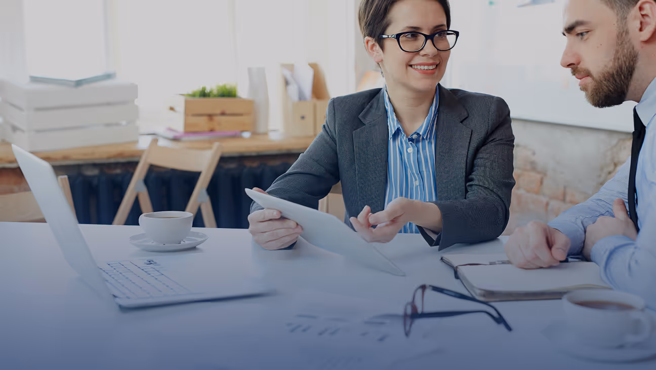 Two business professionals having a discussion at a desk with documents, a laptop, and coffee cups.