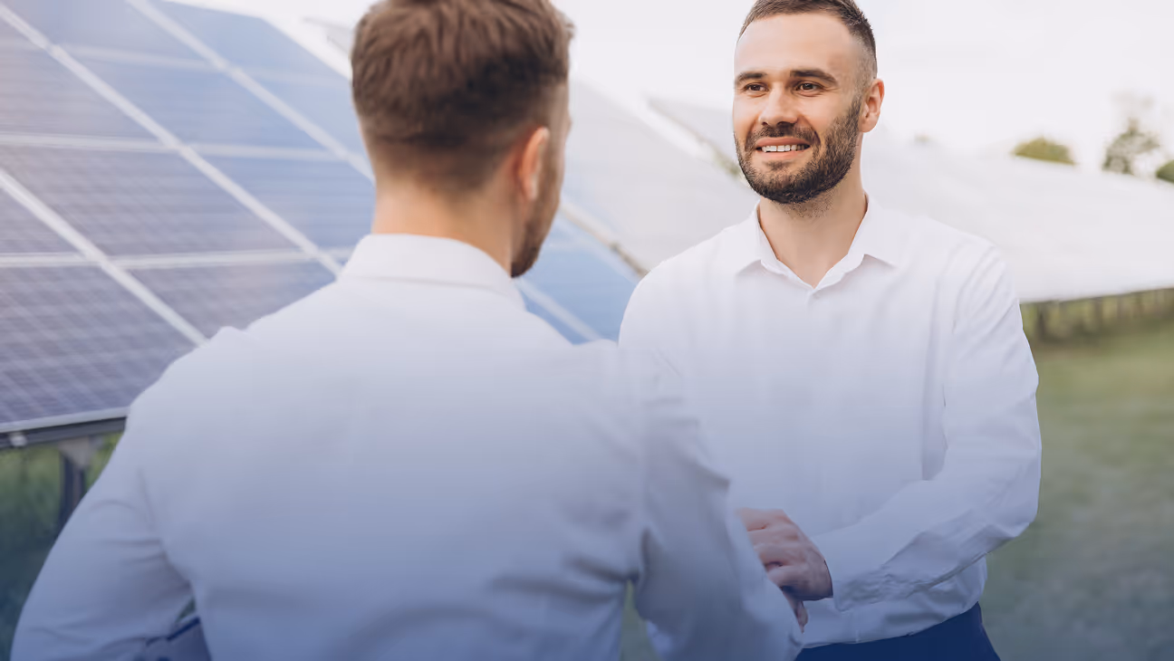 Two men in white shirts shaking hands outdoors near solar panels.