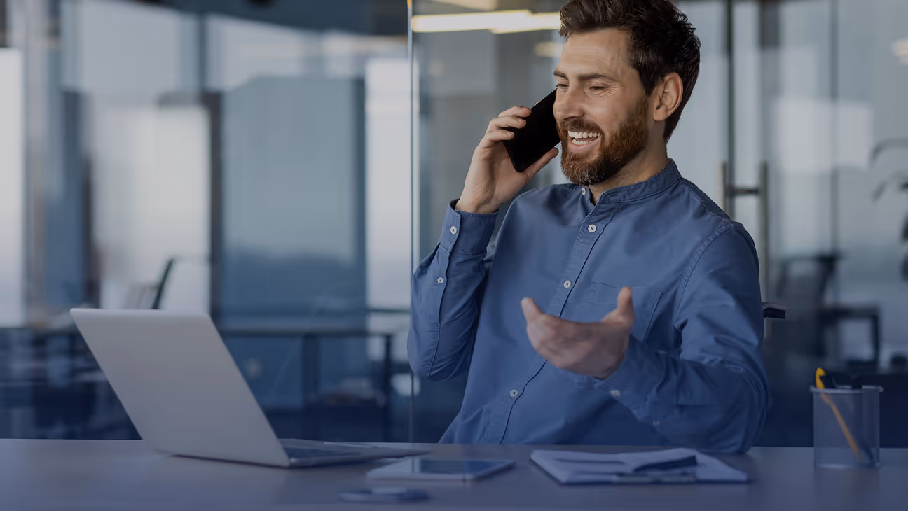 Smiling man in blue shirt talking on a smartphone at a desk with an open laptop and documents.