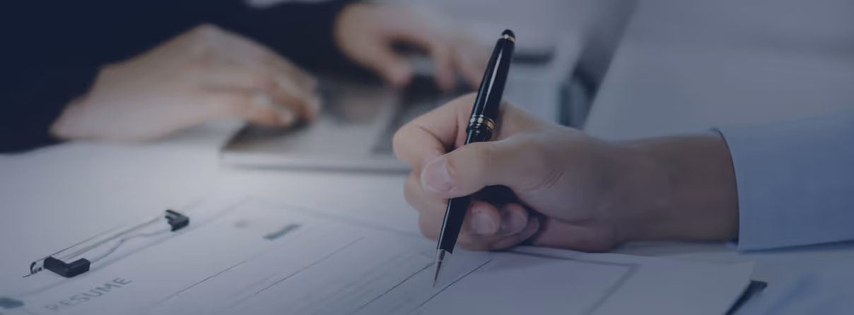 Close-up of a person holding a pen and filling out a resume form on a clipboard, with another person typing on a laptop in the background.