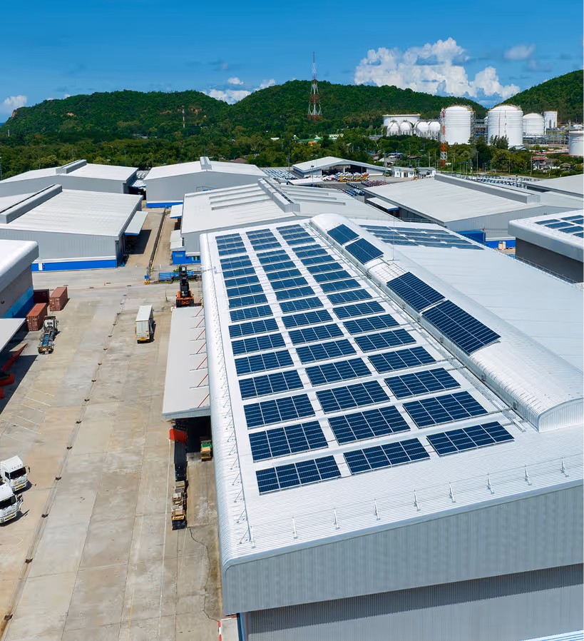 Aerial view of large industrial buildings with solar panels installed on the roof and green hills in the background under a blue sky.
