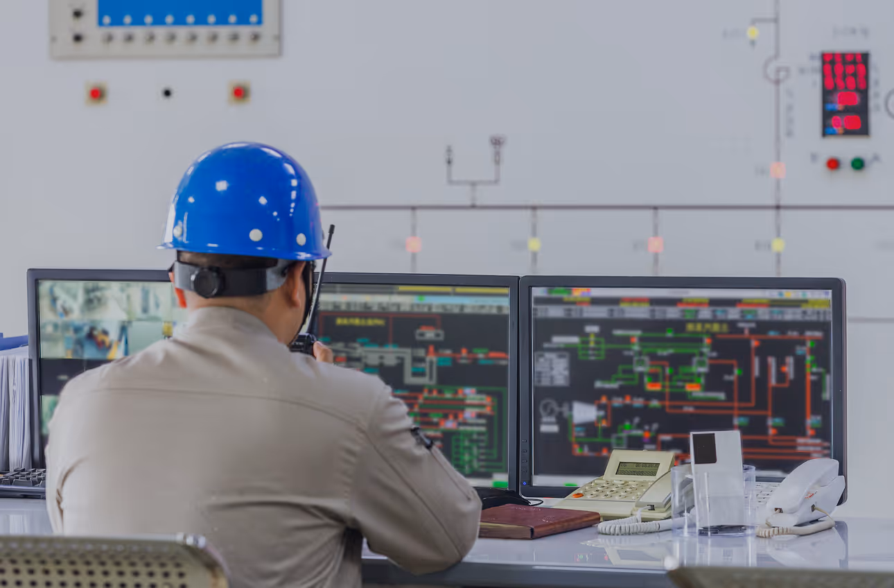 Engineer wearing a blue safety helmet operating a walkie-talkie in front of computer monitors displaying technical schematics.