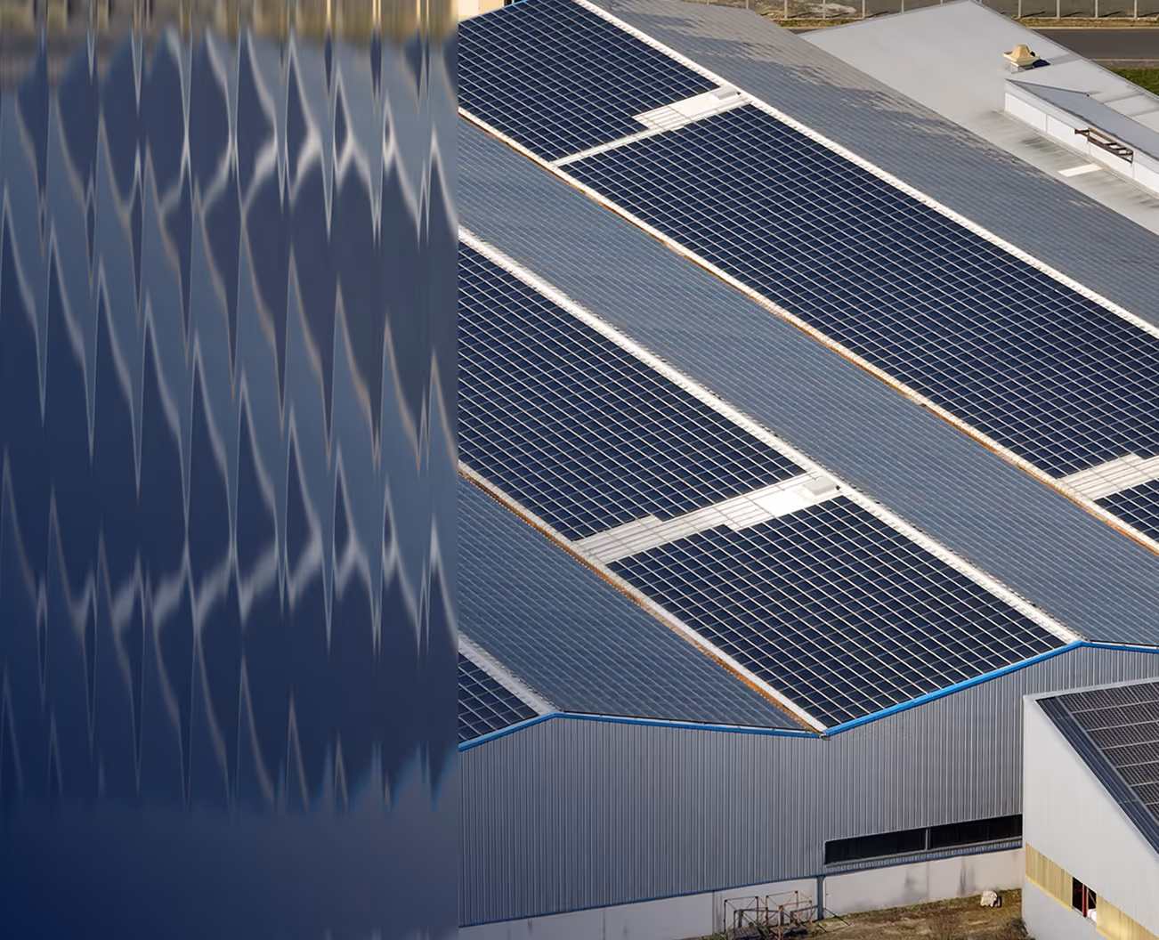 Aerial view of a large industrial building with multiple blue solar panels installed on its roof, reflecting in a nearby mirrored surface.