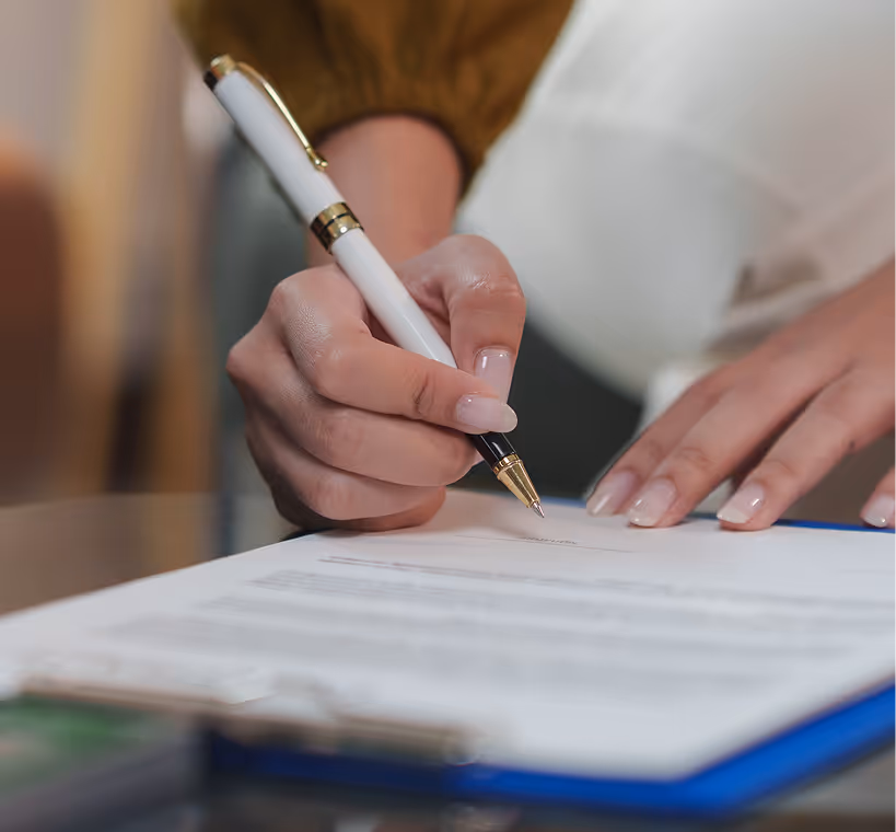 Close-up of a hand holding a white and gold pen signing a document on a table.