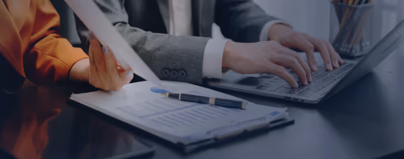 Two people working together at a desk with a laptop and documents, one typing and the other holding papers.