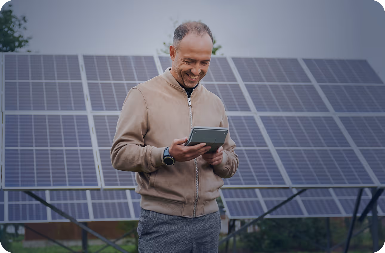 Smiling man in a beige jacket holding a tablet in front of solar panels outdoors.