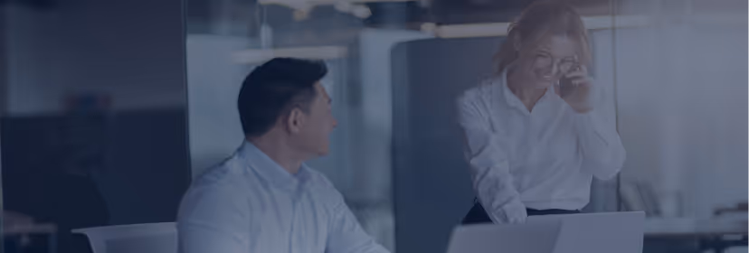 Two business professionals, a man sitting and a woman standing, engaged in conversation in a modern office.