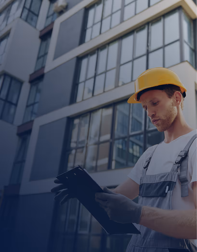 Construction worker in a yellow helmet and gray gloves holding a clipboard in front of a modern glass building.