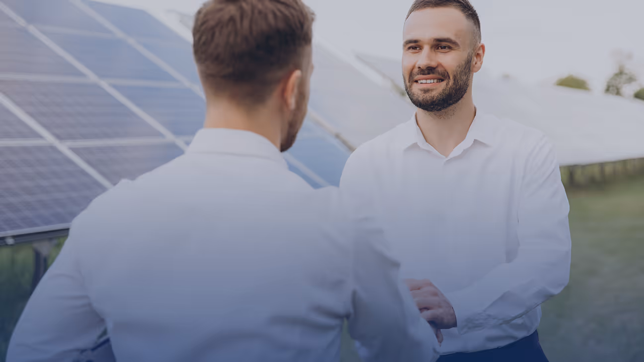 Two men in white shirts shaking hands outdoors near solar panels.