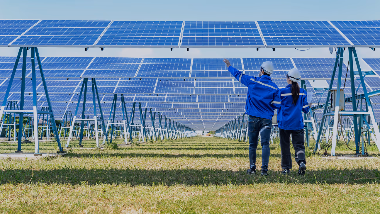 Two engineers in blue jackets and white helmets inspecting a large solar panel array in a grassy field under a clear sky.