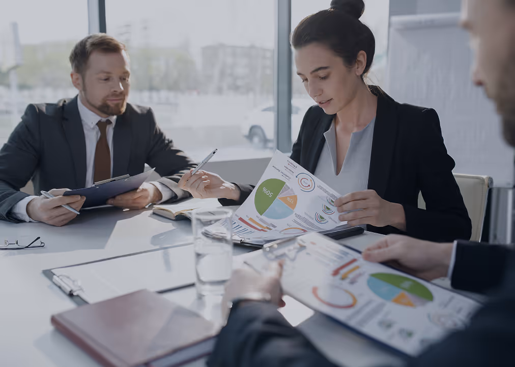 Three business professionals in suits reviewing financial charts during a meeting at a bright office table.