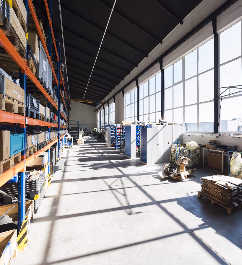 Bright warehouse interior with tall racks stacked with boxes on the left and large windows casting shadows on the floor.