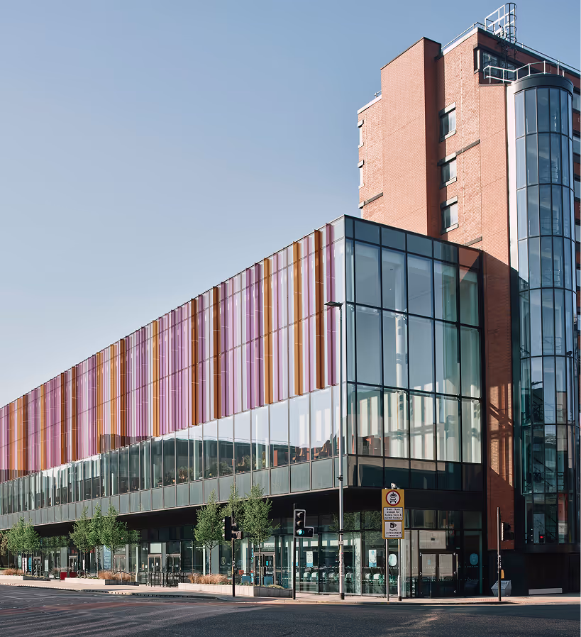 Modern multi-story building with glass walls and colorful vertical stripes on the upper facade, located at a street corner with traffic lights and trees.