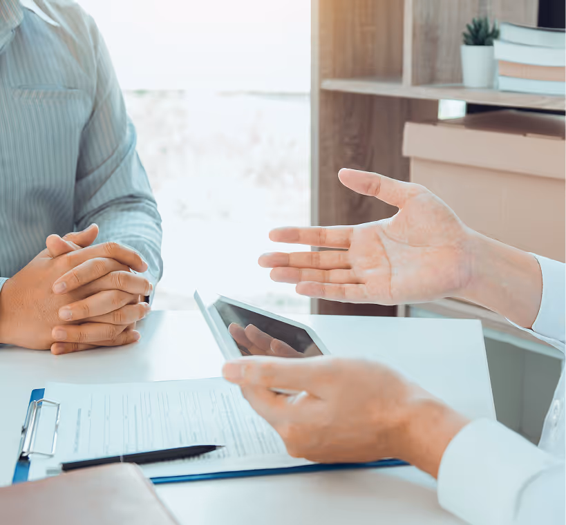 Two people in a discussion at a desk with a tablet and clipboard with documents.