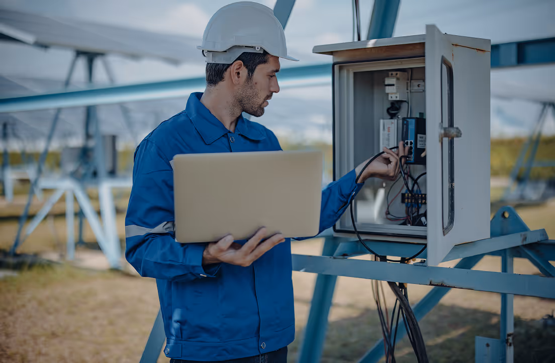 Technician wearing a white hard hat and blue uniform holding a laptop and inspecting an open electrical control panel outdoors.
