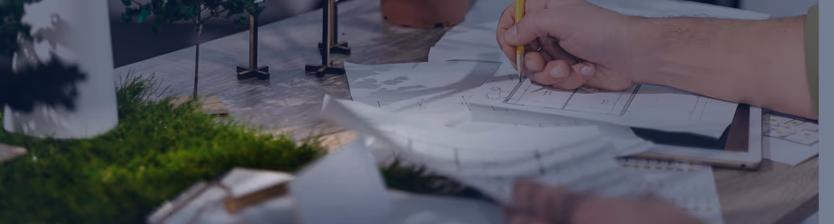 Hand sketching architectural plans on paper surrounded by miniature model trees and green moss.