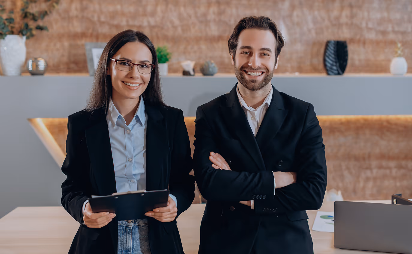 Smiling businesswoman holding a clipboard and businessman with crossed arms standing side by side in a modern office.