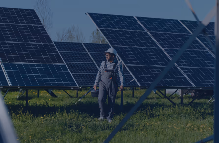 Man in safety helmet and overalls inspecting solar panels in a grassy field on a sunny day.