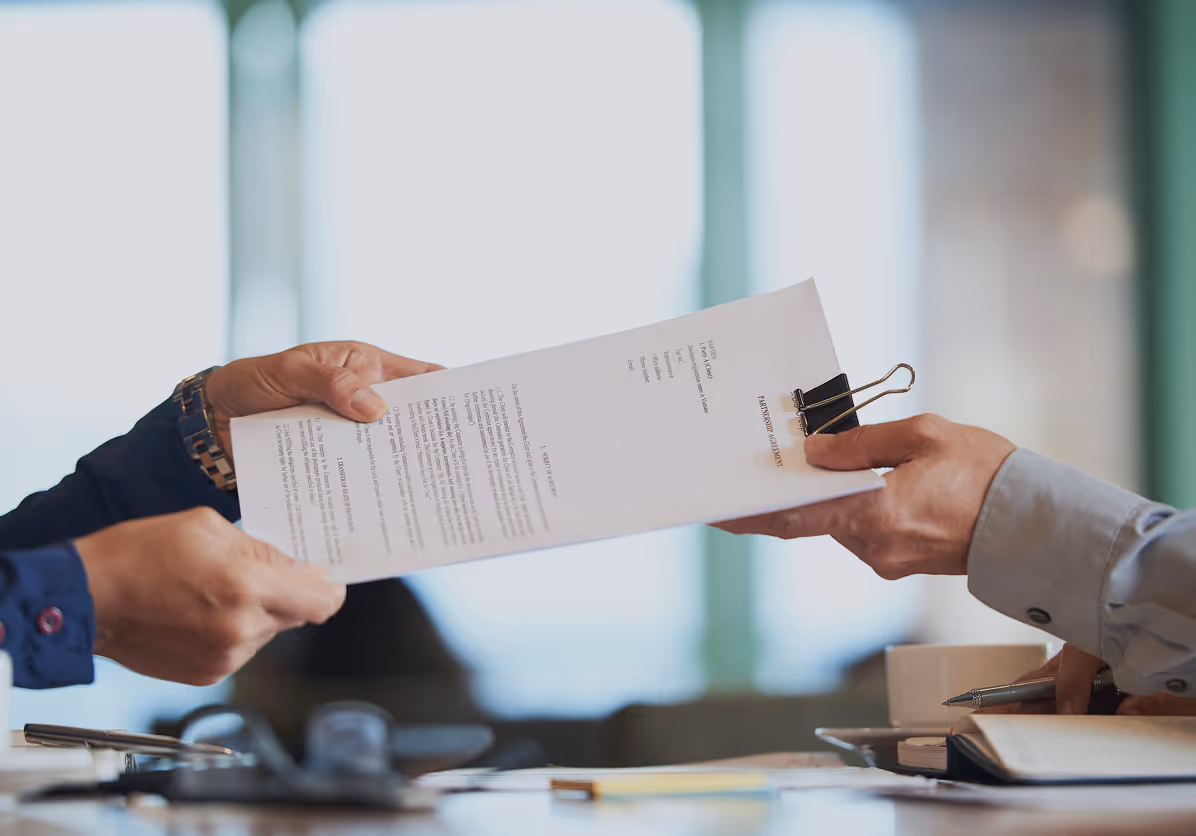 Two people exchanging a document clipped with a black binder in an office setting.
